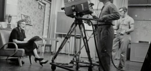 grayscale photography of man standing near studio camera and woman sitting while holding book